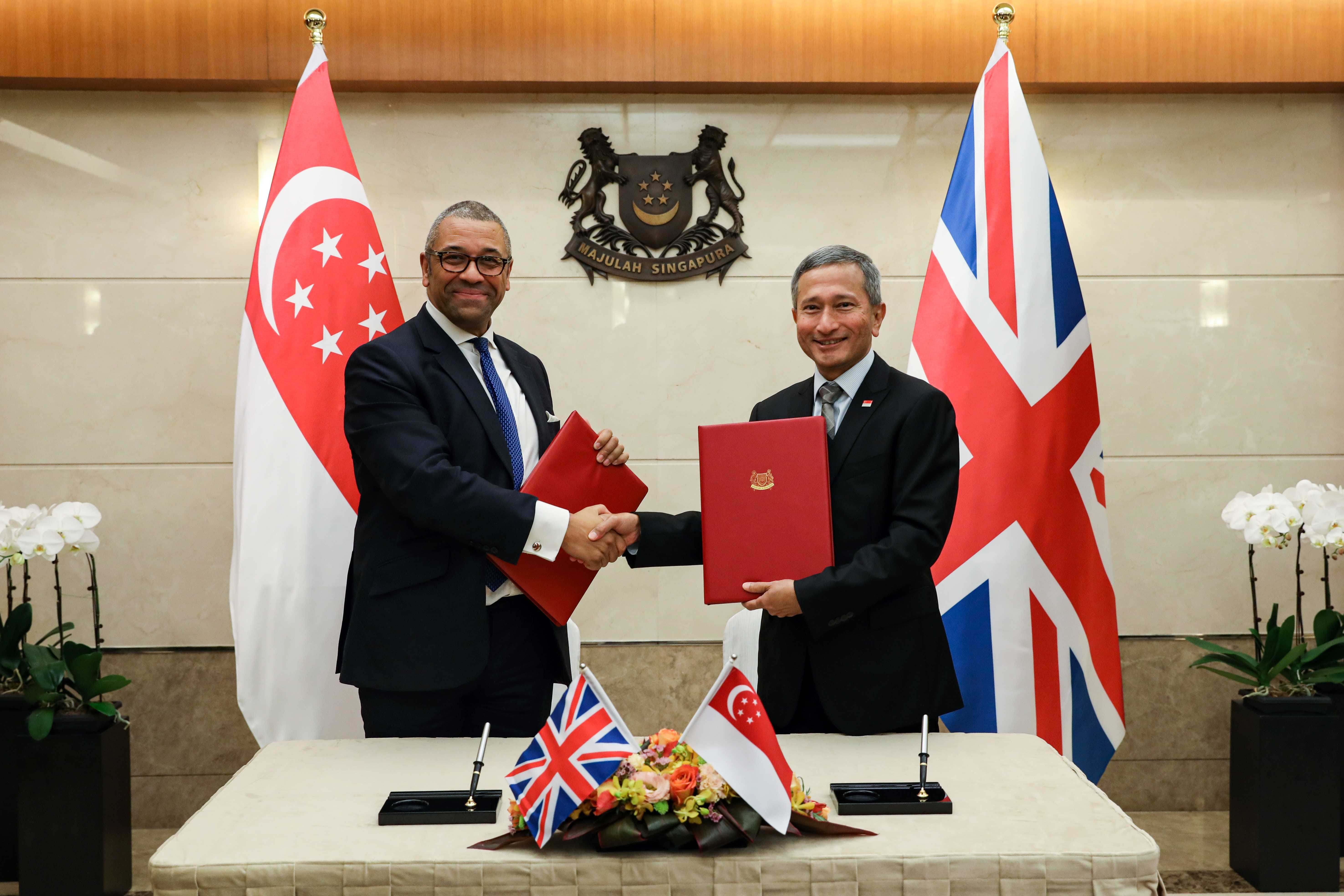 Two men in suits shake hands, holding red folders, flanked by Singapore and UK flags.